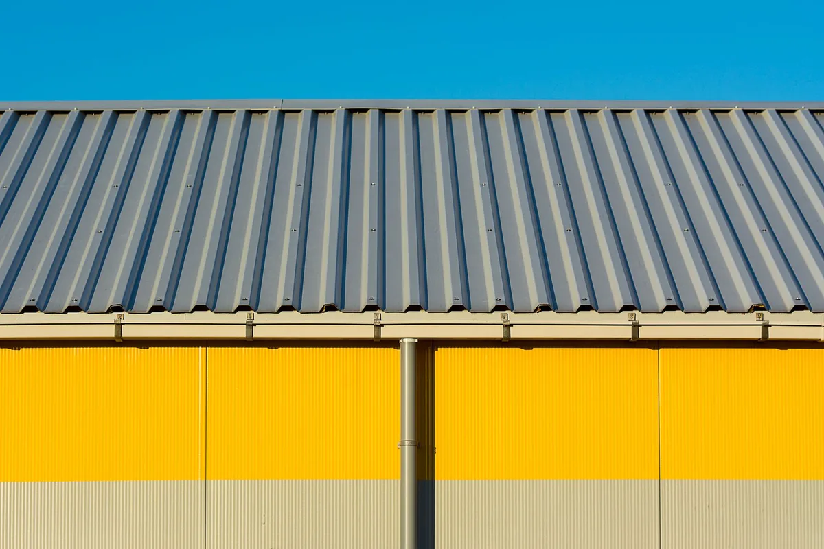 Corrugated metal roof edge with a vertical downpipe running down a yellow wall