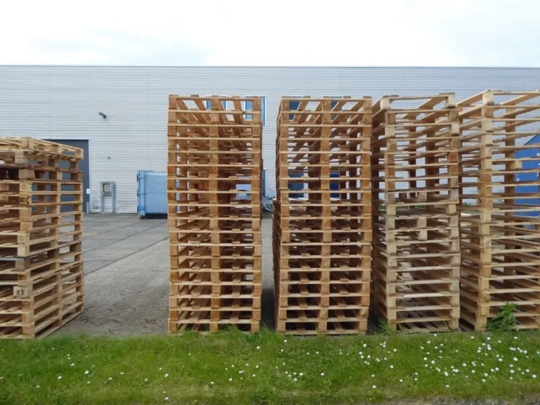 Stacks of wooden pallets on grass beside an industrial warehouse wall